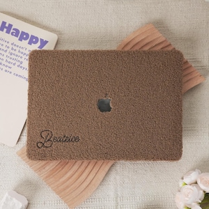 Può includere: Una custodia per laptop marrone e soffice con il logo Apple ritagliato. Il nome "Beatrice" è ricamato in un carattere corsivo. Sullo sfondo è visibile una carta con la parola "Happy".