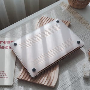 May include: A light pink laptop case with a series of ventilation slots and four black rubber feet. The case is resting on a wooden decorative piece with a wavy design. The laptop case is a protective accessory for a computer.