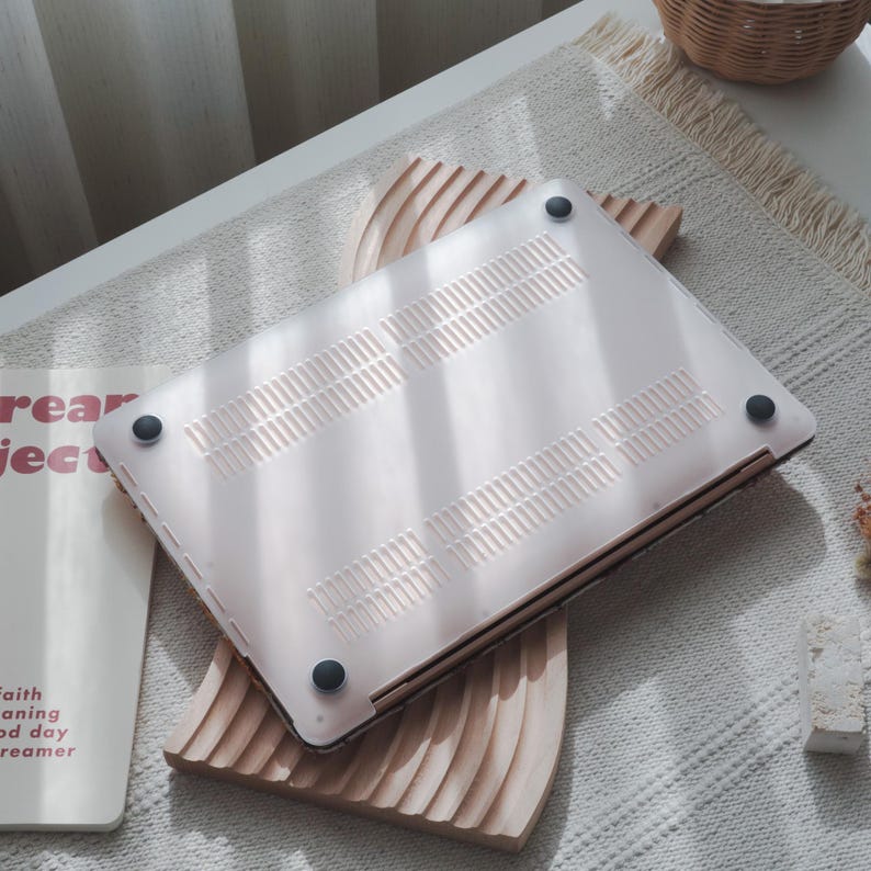 May include: A light pink laptop case with a series of ventilation slots and four black rubber feet. The case is resting on a wooden decorative piece with a wavy design. The laptop case is a protective accessory for a computer.