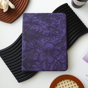 Peut inclure: Un journal violet avec un motif floral. La couverture présente un motif en relief de fleurs d'hibiscus et de feuilles tropicales. Le journal est rectangulaire et semble être fait de cuir ou d'un matériau similaire. Le design est détaillé et complexe.