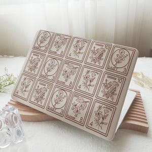 Könnte beinhalten: Eine beige Laptop-Hülle mit einem sich wiederholenden Muster aus braunen Blumenmotiven im Poststempel-Stil. Jeder Stempel enthält eine Blumenillustration, Zahlen und die Wörter "DAISY", "FLOWER" und "JASMINE".