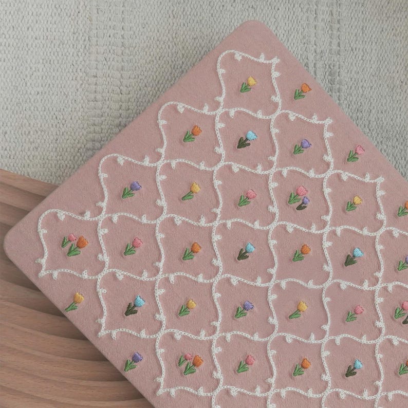 May include: A pink, square-shaped textile with a repeating pattern of embroidered tulips in various colors. The tulips are framed by a white, decorative border. The textile is placed on a wooden surface.