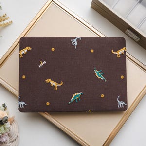 Op de afbeelding: Een rechthoekige, bruine etui met geborduurde dinosaurusontwerpen in geel, blauw en wit. De etui is versierd met kleine gele stervormige accenten. De etui ligt op een goudomrand oppervlak.
