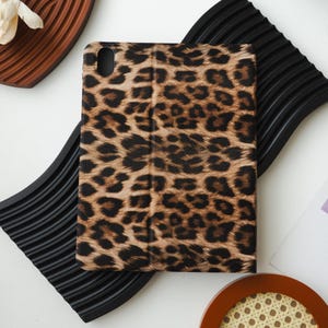 Puede incluir: Una funda para tableta con un diseño de estampado de leopardo en tonos marrones y negros. La funda está cerrada y tiene un acabado liso y brillante. La funda es rectangular y parece estar hecha de un material duradero.