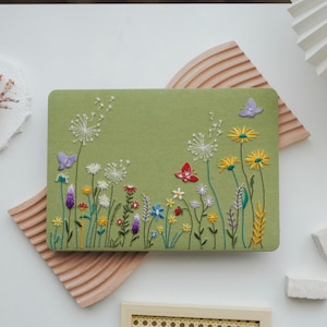 Puede incluir: Una funda rectangular verde oliva con un diseño floral bordado. El bordado incluye dientes de león blancos, margaritas amarillas y flores silvestres coloridas, con tres mariposas. La funda es probablemente para un portátil o una tableta.
