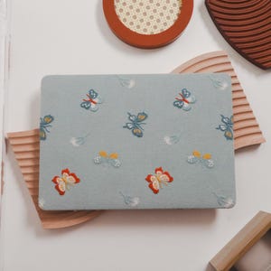 Könnte beinhalten: Eine hellblaue, rechteckige Laptop-Hülle mit gestickten Schmetterlingen in Rot, Gelb und Blau. Die Schmetterlinge sind über die Oberfläche verstreut, mit kleinen weißen floralen Akzenten. Die Hülle liegt auf einer Holzoberfläche.