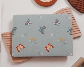 May include: A light blue rectangular laptop case with embroidered butterflies in red, yellow, and blue. The butterflies are scattered across the surface, with small white floral accents. The case is resting on a wooden surface.