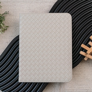 May include: A light gray, woven-patterned notebook or journal cover. The cover has a rectangular shape with rounded corners and a stitched edge. The background includes a black, wavy-edged tray and a neutral-colored textile.