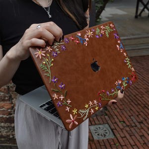 May include: A brown laptop case featuring an embroidered floral pattern with small pink, purple, yellow, and blue flowers and green stems. The case has a cutout for the Apple logo and covers the lid of a silver laptop with a black keyboard.
