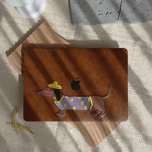 Peut inclure: Une coque d'ordinateur portable marron avec un teckel brodé portant un chapeau jaune et un pull violet à pois jaunes. L'étui a une texture semblable à du cuir marron et un logo Apple noir.