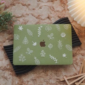 Könnte beinhalten: Eine grüne Laptop-Hülle mit weißen, gestickten botanischen Designs, darunter Blätter und Blumen. Die Hülle hat ein braunes Apple-Logo und liegt auf einem schwarzen Ständer, vor einem strukturierten, hellbraunen Hintergrund.