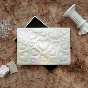 May include: A pearlescent white rectangular case with a heart pattern. The case is open, revealing a black interior. The hearts are of varying sizes and have a stitched outline. The case sits on a textured, brown surface.