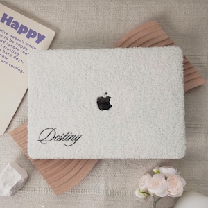 Peut inclure: Une housse d'ordinateur portable blanche et duveteuse avec le logo Apple et le nom "Destiny" en écriture cursive. L'étui repose sur un élément décoratif en bois et une surface beige. Une carte avec le mot "Happy" est visible.