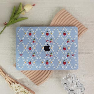 Può includere: Una custodia per laptop azzurra con un motivo a fragole ricamato. La custodia presenta un motivo ripetuto di fragole, fiocchi ed elementi floreali. Il logo Apple è visibile al centro.