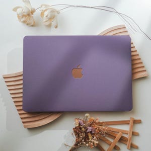 Könnte beinhalten: Ein lila Laptop mit goldenem Apple-Logo liegt auf einer weißen Oberfläche. Er ist von dekorativen Holzelementen und Trockenblumen umgeben. Das Design ist minimalistisch und modern. Abmessungen: ca. 30 cm x 21 cm.