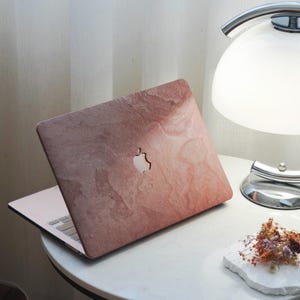 Può includere: Un laptop con un design rosa e marrone simile al marmo sul coperchio, con il logo Apple. Il laptop è aperto, rivelando la tastiera. È appoggiato su un tavolo bianco accanto a una lampada e a una piccola composizione floreale.