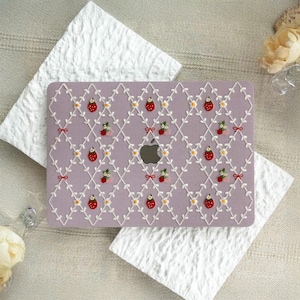 Può includere: Una custodia per laptop lavanda con un ricamo a fragole. La custodia presenta un motivo ripetuto di fragole, fiori bianchi e foglie verdi, collegati da un reticolo bianco. Piccoli fiocchi rossi accentuano il design. Il logo Apple è visibile.