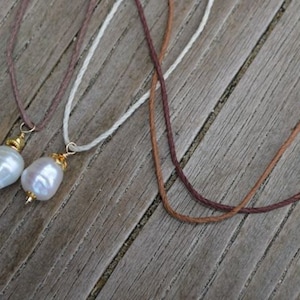 May include: Three necklaces with natural pearls and cord are displayed. Two necklaces have large, irregular white pearls with gold-coloured accents. The cords are brown, white, and dark brown. The background is a weathered wooden surface.