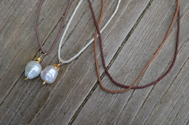 May include: Three necklaces with natural pearls and cord are displayed. Two necklaces have large, irregular white pearls with gold-coloured accents. The cords are brown, white, and dark brown. The background is a weathered wooden surface.