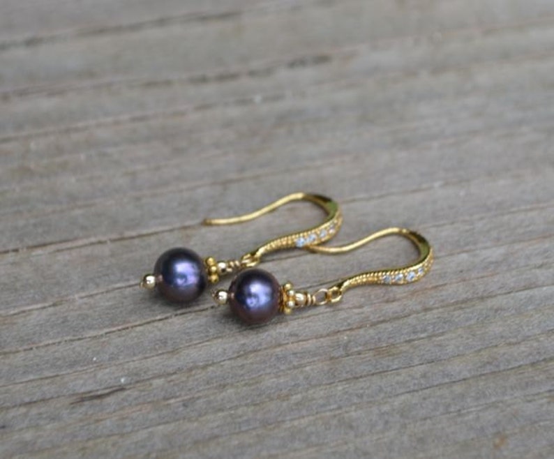 May include: A pair of gold-coloured earrings featuring dark grey pearls and small clear stones. The earrings have a curved design with a hook closure. The pearls are round and have an iridescent sheen.