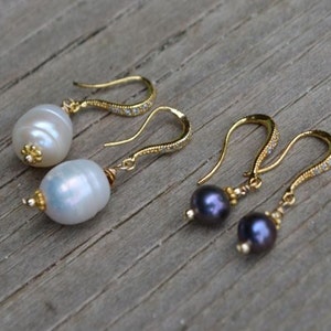 May include: Two pairs of drop earrings with gold-coloured hooks. One pair features large, white, oval pearls, the other has dark purple, round pearls. The hooks are adorned with small, clear crystals.