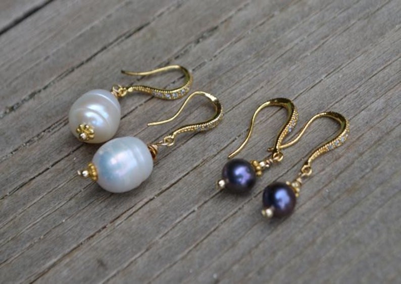 May include: Two pairs of drop earrings with gold-coloured hooks. One pair features large, white, oval pearls, the other has dark purple, round pearls. The hooks are adorned with small, clear crystals.