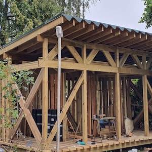 Op de afbeelding: Een houten frameconstructie met een groen dak, in aanbouw. Het frame is gemaakt van onbehandeld hout en wordt ondersteund door houten balken. Het dak is gemaakt van gegolfd metaal.