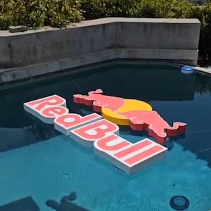 May include: Large, floating Red Bull logo and bull graphic in a swimming pool. The logo is white with red lettering and a red outline. The bull graphic is red with a yellow sun. The pool has blue water.