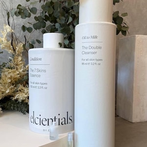 May include: White cylindrical skincare product bottles with black text, including "Oil to Milk The Double Cleanser" and "The 7 Skins Essence." The bottles are displayed on a white platform with greenery in the background. Also included are two small sample bottles.