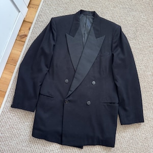 Może przedstawiać: Czarny dwurzędowy blazer z satynowymi klapami. Blazer ma dwa rzędy guzików i dwie przednie kieszenie. Rękawy mają guziki przy mankietach. Blazer leży płasko na dywanie w neutralnym kolorze.