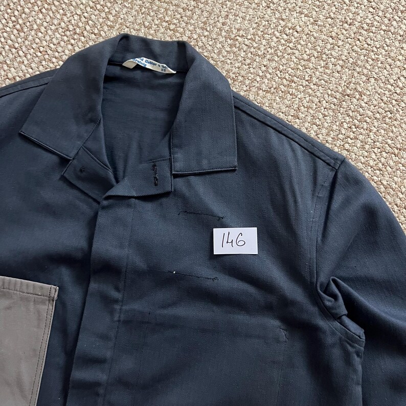 Puede incluir: Una camisa de trabajo azul marino oscuro con cuello y cierre de botones. La camisa tiene una peque&ntilde;a etiqueta blanca con el n&uacute;mero "146" escrito. Un bolsillo beige es visible en la parte inferior izquierda.