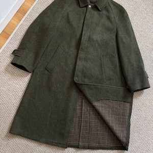 May include: An olive green wool coat featuring a classic collar, a concealed button placket, and raglan sleeves. The coat has two side pockets and adjustable button tabs on the cuffs. The interior lining displays a brown and black houndstooth pattern.