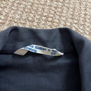 Puede incluir: Primer plano de un cuello de prenda azul oscuro con una etiqueta blanca. La etiqueta dice "Gebler & Son Breinig 52". La tela tiene un sutil patr&oacute;n de espiga.