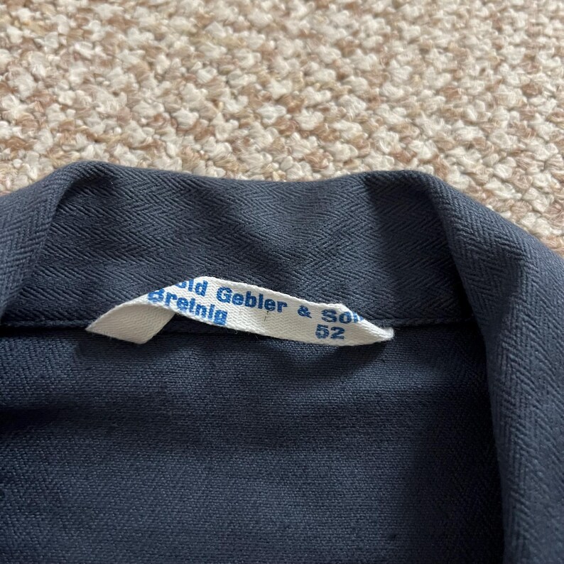 Puede incluir: Primer plano de un cuello de prenda azul oscuro con una etiqueta blanca. La etiqueta dice "Gebler & Son Breinig 52". La tela tiene un sutil patr&oacute;n de espiga.