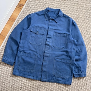 May include: A blue work jacket with four front pockets and a button-down closure. The jacket has a collared neckline and long sleeves. The fabric appears to be a durable cotton or similar material, suitable for workwear. The jacket is laid flat on a neutral-toned rug.