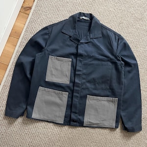 Puede incluir: Chaqueta de trabajo azul marino con cuatro bolsillos cuadrados grises. La chaqueta tiene cuello y cierre de botones. La prenda est&aacute; hecha de una tela resistente y est&aacute; colocada sobre una alfombra de color neutro.