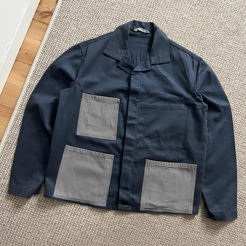 Puede incluir: Chaqueta de trabajo azul marino con cuatro bolsillos cuadrados grises. La chaqueta tiene cuello y cierre de botones. La prenda est&aacute; hecha de una tela resistente y est&aacute; colocada sobre una alfombra de color neutro.