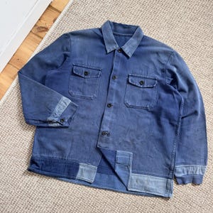 Puede incluir: Chaqueta de trabajo azul vintage con dos bolsillos en el pecho y cierres de botones. La chaqueta tiene cuello y puños abotonados. La tela muestra signos de desgaste, lo que aumenta su atractivo vintage. La chaqueta es de color azul medio.