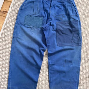 以下が含まれることがあります： 2つの大きなパッチポケットと左脚のデニムパッチが付いた青いワークパンツ。パンツは、フロントにギャザーの入った伸縮性のあるウエストバンドとプリーツを備えたリラックスフィットです。パンツは無地の青色です。