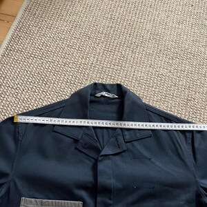 Puede incluir: Una camisa de trabajo azul oscuro con una cinta m&eacute;trica colocada sobre el cuello. La cinta m&eacute;trica es blanca con marcas negras, que muestran medidas en cent&iacute;metros. La camisa tiene cuello y un bolsillo en el lado inferior izquierdo.