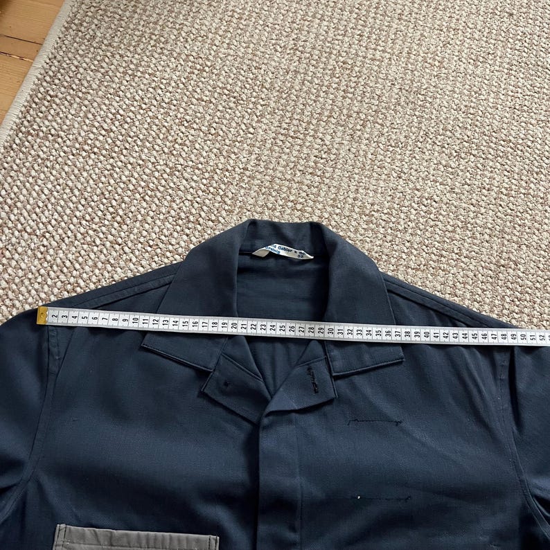 Puede incluir: Una camisa de trabajo azul oscuro con una cinta m&eacute;trica colocada sobre el cuello. La cinta m&eacute;trica es blanca con marcas negras, que muestran medidas en cent&iacute;metros. La camisa tiene cuello y un bolsillo en el lado inferior izquierdo.
