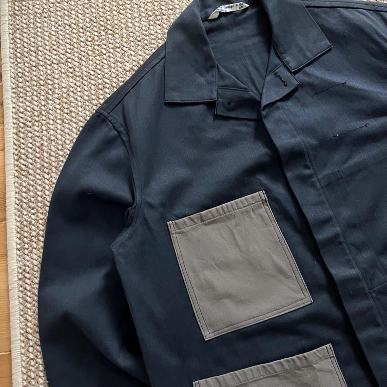 Puede incluir: Chaqueta de trabajo azul marino con dos grandes bolsillos cuadrados grises en la parte delantera. La chaqueta tiene cuello y cierre de botones. La prenda est&aacute; hecha de una tela duradera, adecuada para el trabajo o el uso diario.