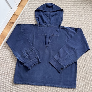 May include: A navy blue hooded shirt with long sleeves. The shirt has a v-neck opening and buttoned cuffs. The fabric appears to be a sturdy cotton or similar material. The garment is laid flat on a neutral-colored surface.