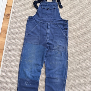 Può includere: Salopette in denim blu con spalline blu scuro e multiple tasche. L'indumento da lavoro mostra segni di usura, con macchie blu più chiare e schizzi di vernice. La salopette è stesa su un tappeto di colore neutro.