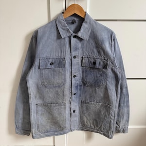 Può includere: Giacca da lavoro blu sbiadita con quattro tasche frontali e chiusura con bottoni. La giacca è appesa a una gruccia di legno. La giacca ha un aspetto usurato e vintage. Lo sfondo è bianco.