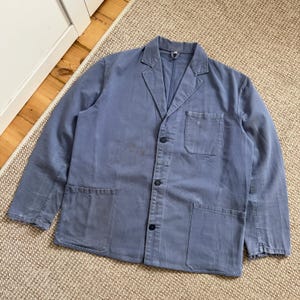 May include: A dusty blue jacket with a classic blazer design. The jacket features a notched collar, three buttons, and three pockets. The fabric shows signs of wear and tear, adding to its vintage appeal. The jacket is laid flat on a neutral-toned rug.
