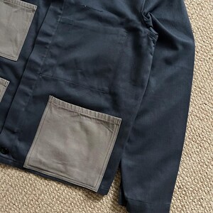 Puede incluir: Chaqueta de trabajo azul marino con tres bolsillos grandes, cuadrados y grises. La chaqueta tiene cierre de botones y un dise&ntilde;o sencillo y utilitario. La tela parece ser un material duradero de peso medio, adecuado para el trabajo o el uso informal.