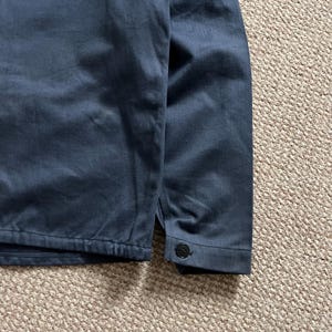 Puede incluir: Primer plano de una chaqueta azul marino. La chaqueta tiene un pu&ntilde;o abotonado y un cierre de bot&oacute;n en el dobladillo. La tela parece ser un algod&oacute;n o mezcla resistente.