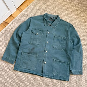 Puede incluir: Una chaqueta de trabajo de manga larga en color verde azulado oscuro con cuello clásico, cierre frontal de botones metálicos plateados, dos bolsillos en el pecho con solapa y dos grandes bolsillos de parche en la parte inferior frontal.