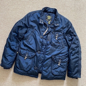 Könnte beinhalten: Eine marineblaue Steppjacke mit mehreren Taschen und einem Reißverschluss. Die Jacke hat ein gelbes und schwarzes Etikett mit dem Text "CP Company" auf der linken Brustseite.